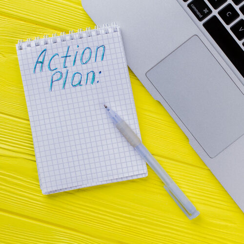 Notepad with action plan and laptop pc. Flat lay top view. Yellow wooden background.
